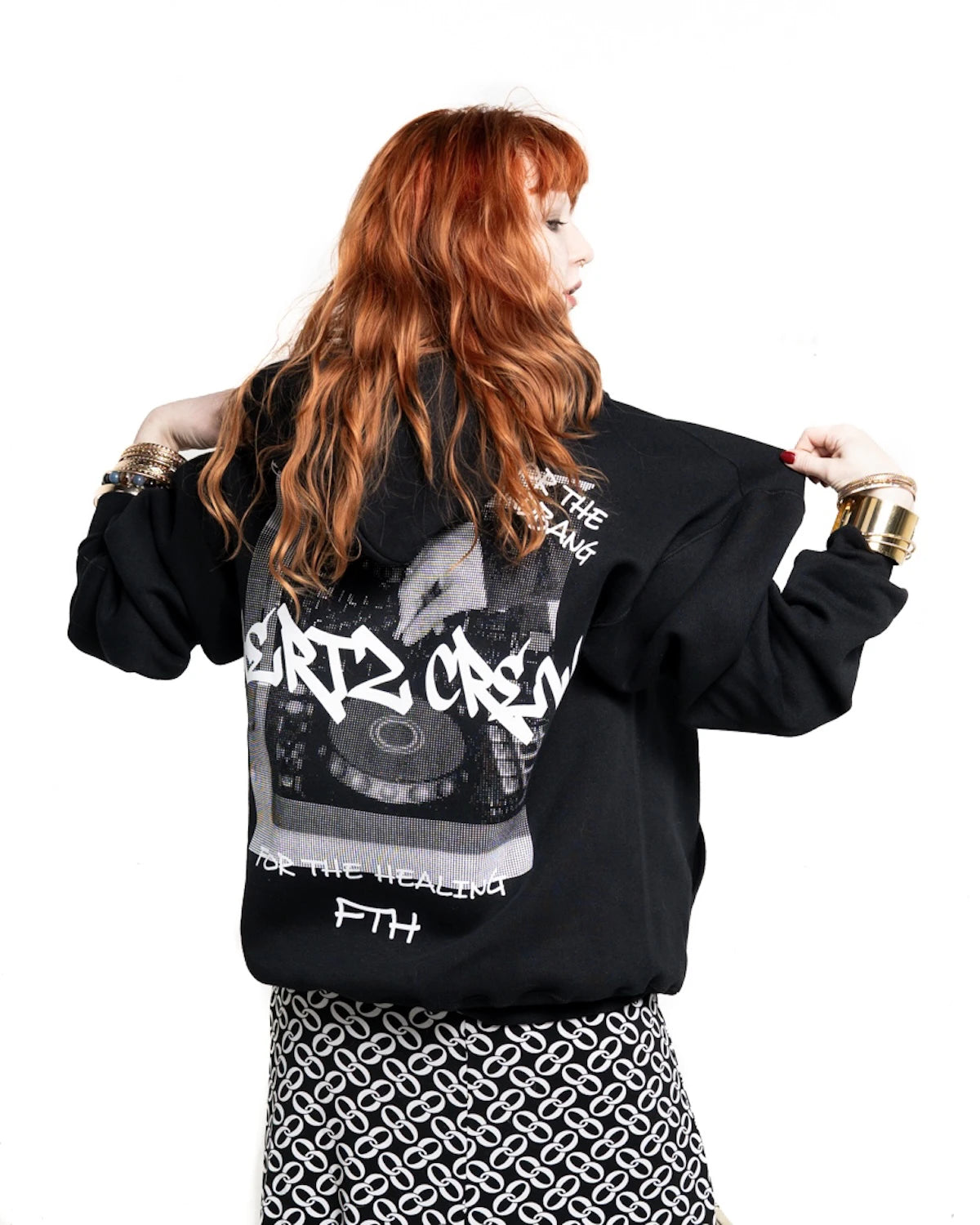 Red headed woman wearing a black hooded sweatshirt with hertz crew in graffiti font with halftone of hand playing dj controller